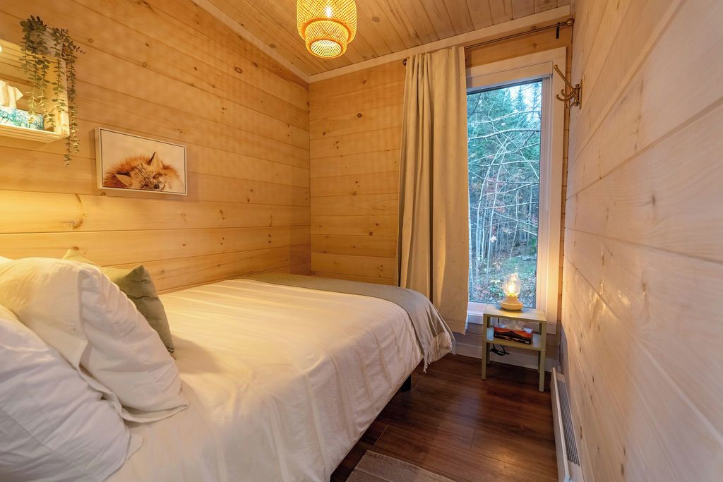Un dormitorio de cabaña de madera con una cama, un cuadro enmarcado de un zorro, una planta colgante y una ventana con cortinas.