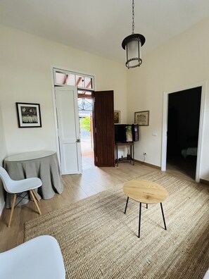 A living room with a round coffee table, two white chairs, a small side table, a hanging light fixture, and a door leading to a balcony.