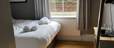 A bedroom with a bed, a window with curtains, a wooden floor, and a desk with a lamp.