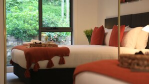 Ein ordentlich bezogenes Bett mit roter Decke und Kissen, ein Nachttisch mit einer Snackauswahl und Blick ins Grüne vor dem Fenster.