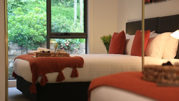 Ein ordentlich bezogenes Bett mit roter Decke und Kissen, ein Nachttisch mit einer Snackauswahl und Blick ins Grüne vor dem Fenster.