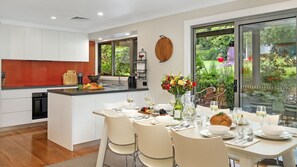 Una cocina moderna con armarios blancos, un salpicadero rojo y una isla grande con un lavamanos. Hay una zona de comedor con una mesa preparada para seis personas, rodeada de sillas.