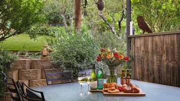 Una mesa de vidrio con una botella de vino, una copa de vino blanco, una botella de cerveza, un florero con flores y una tabla de madera con pasteles y frutas.