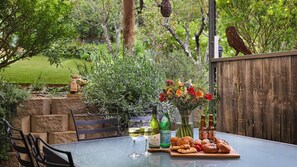 Une table en verre sur laquelle se trouvent une bouteille de vin, un verre de vin blanc, une bouteille de bière, un vase de fleurs et une planche en bois avec des pâtisseries et des fruits.
