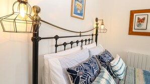 En säng med en svart metallram, vita sängkläder och mönstrade kuddar. På väggen sitter en lampa med en synlig glödlampa.