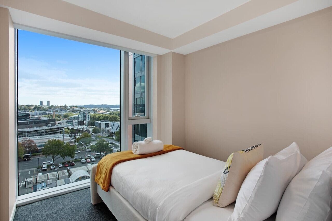 Een hotelkamer met een groot raam met uitzicht op de stad, een net opgemaakt bed met witte lakens en een bruine plaid.