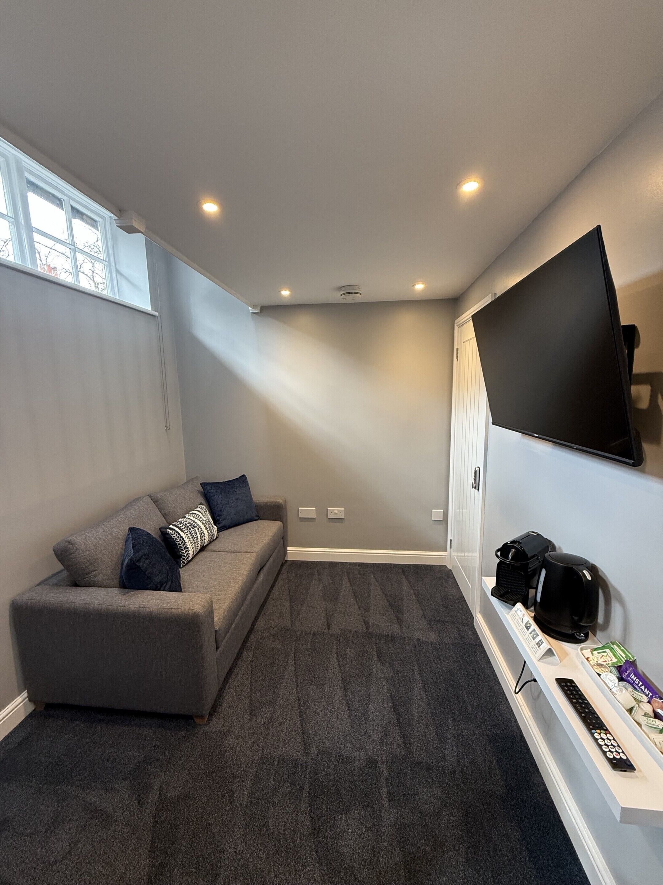 A modern living room with a grey sofa, a flat-screen TV mounted on the wall, and a coffee maker on a side table.