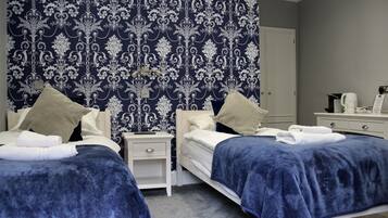 A hotel room with two beds, each with a blue blanket and pillows, a nightstand with a lamp, and a dresser with a coffee maker.