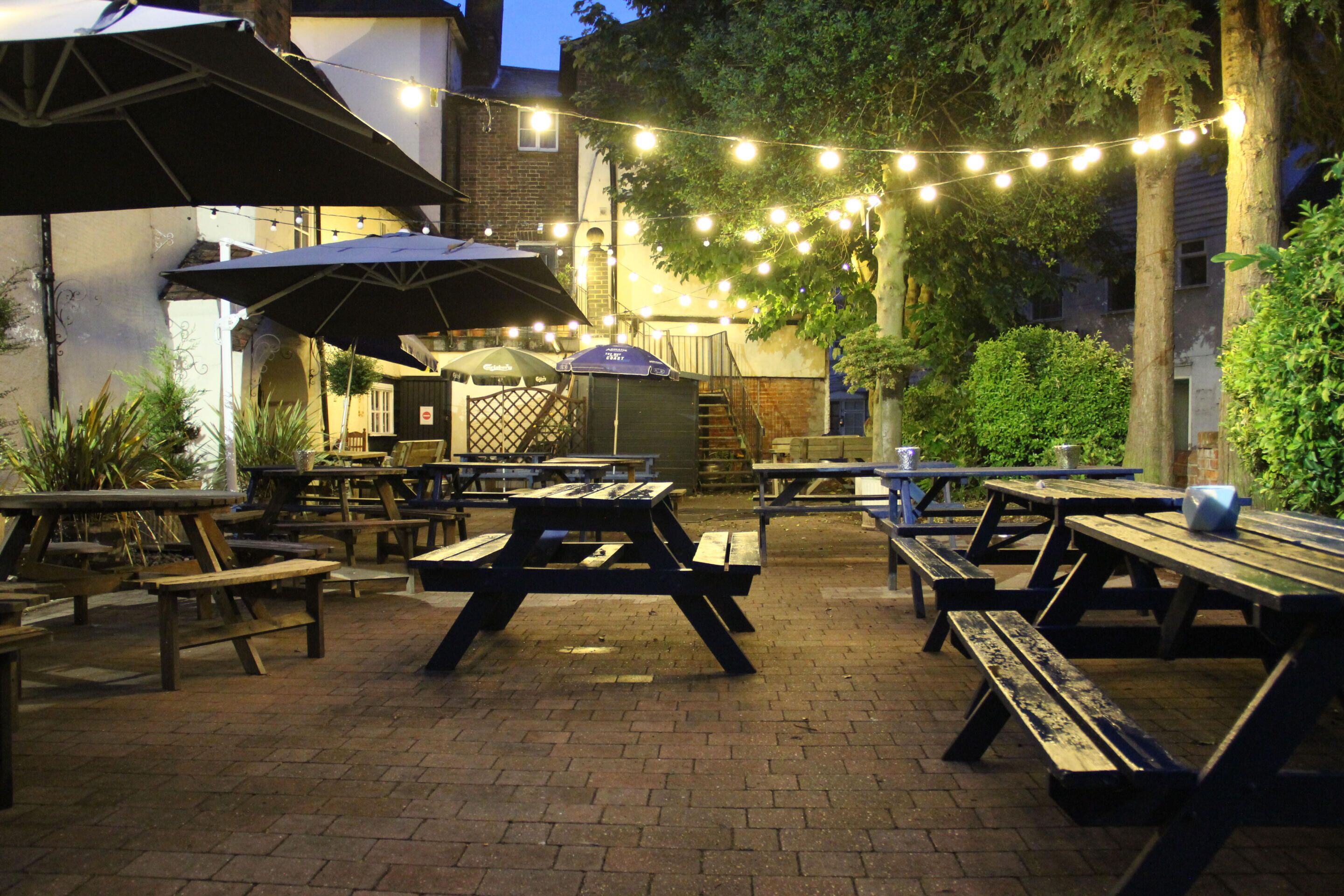 An outdoor patio area with picnic tables and umbrellas, string lights, and a barbecue grill.