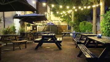 An outdoor patio area with picnic tables and umbrellas, string lights, and a barbecue grill.