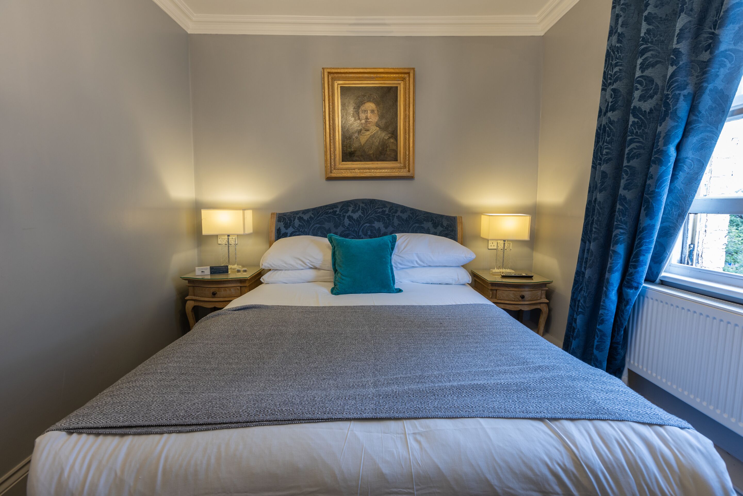 Een opgemaakt bed met een blauw hoofdeinde, twee bedlampjes en een ingelijst portret erboven.