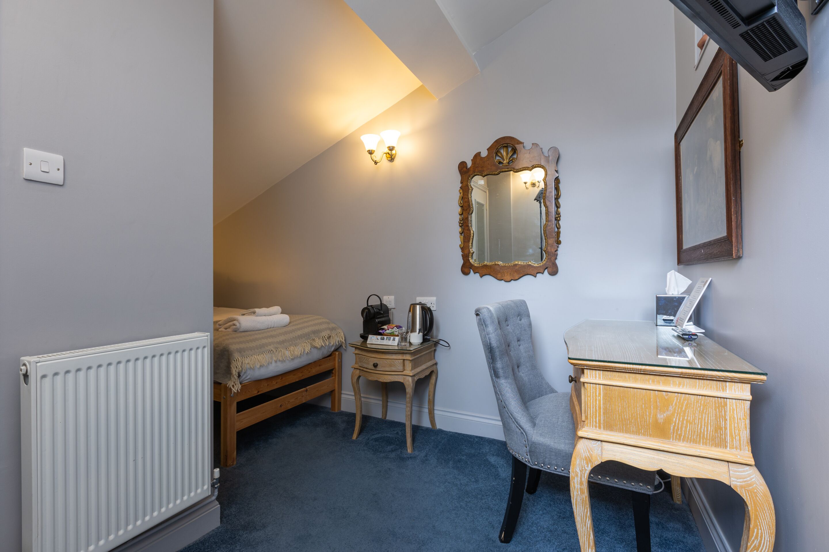 A small bedroom with a bed, a wooden nightstand, a chair, a desk with a laptop, a mirror, and a radiator.