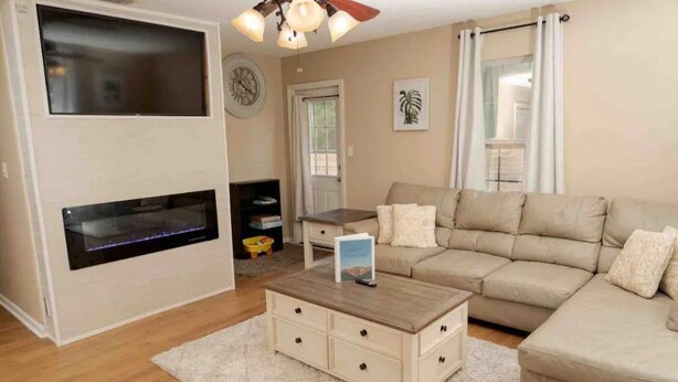 Una sala de estar con un sofá color crema, una mesa de centro, un televisor de pantalla plana montado en la pared y un ventilador de techo.