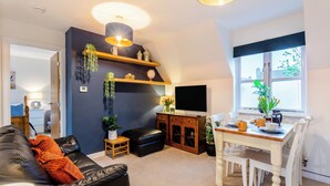 A modern living room with a black leather sofa, a dining table with chairs, a TV mounted on the wall, and a large window providing natural light. - Delightful recently completed Annexe close to Cambridge. (Cambridge)
