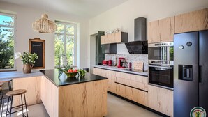 A modern kitchen with wooden cabinetry, a central island, a black refrigerator, and a coffee station. - LA Maison DE Ninette (Castelculier)