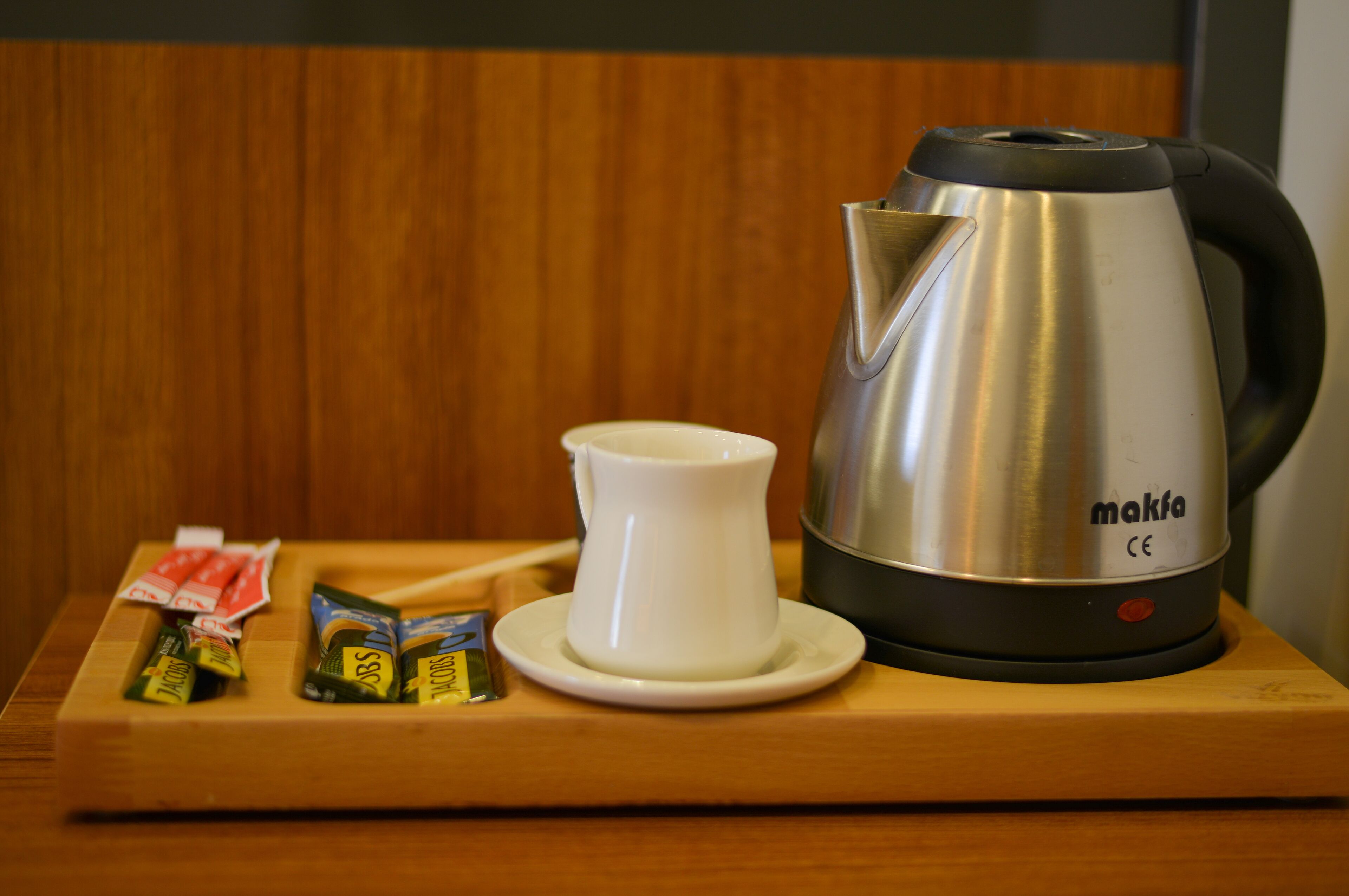 A stainless steel electric kettle with a white cup and saucer, and packets of tea on a wooden tray.