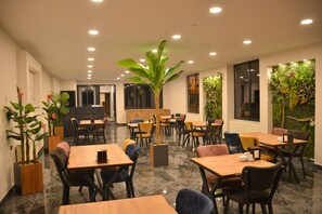 A restaurant with wooden tables and chairs, potted plants, and a large tree in the center.