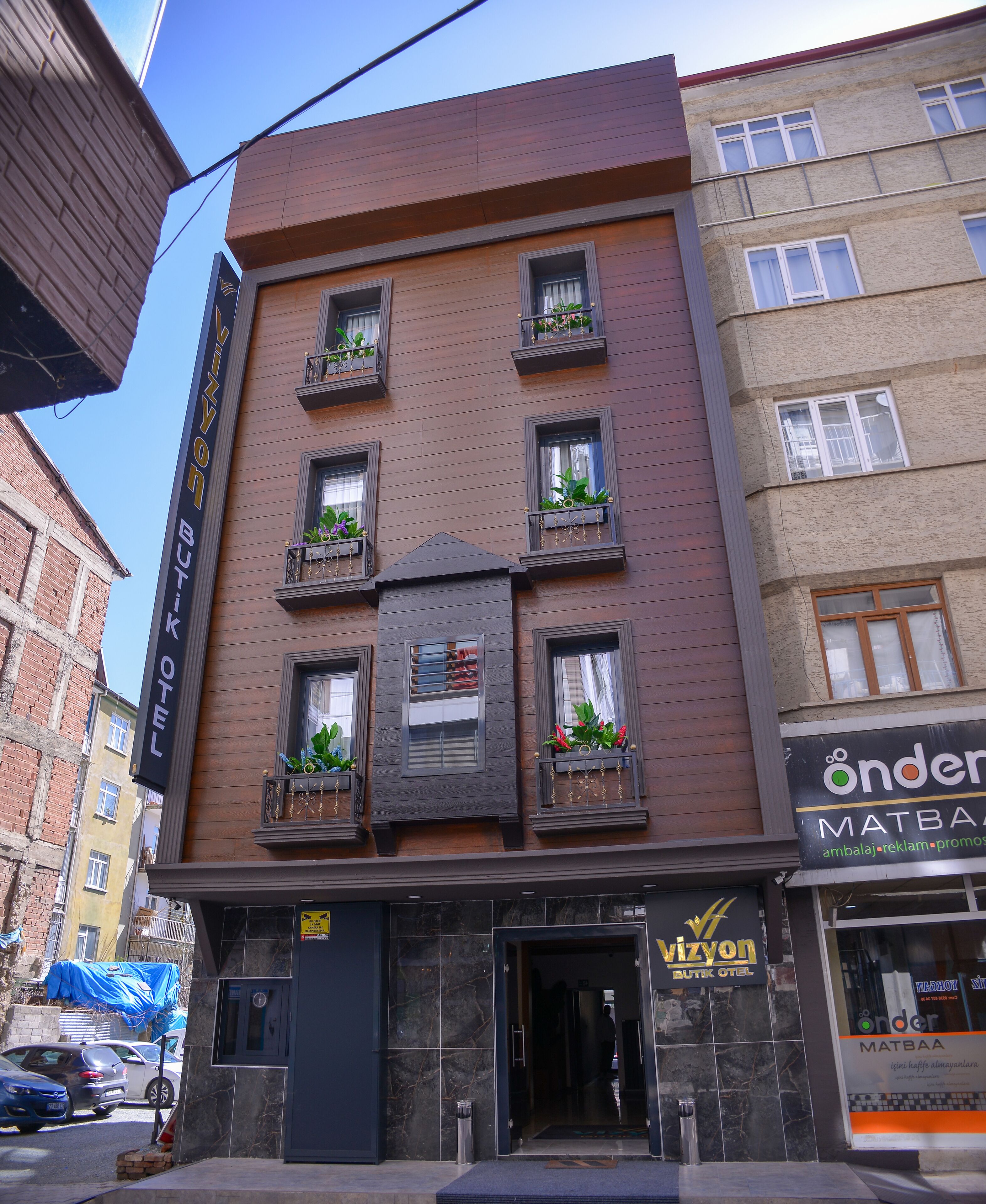 A hotel with a dark brown facade, multiple windows with plants, and a visible entrance.