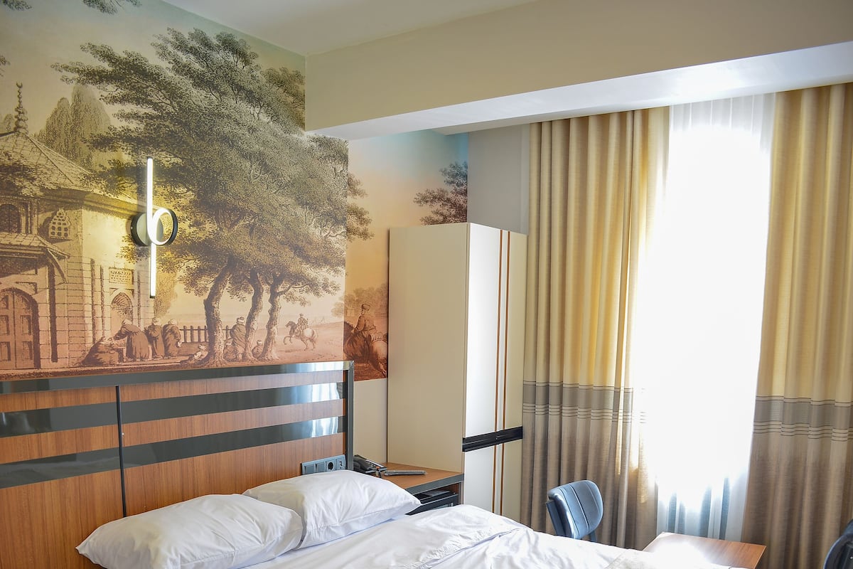 Una cama bien hecha con sábanas blancas y un plaid color beige, un cabecero de madera, una lámpara de pared y un ventanal con cortinas.