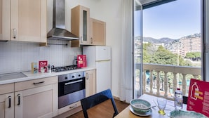 A modern kitchen with wooden cabinets, a gas stove, and an oven. A dining table with a chair is visible, and a large window offers a view of a cityscape and hills. - Casa Camilla al Mare by Wonderful Italy (Genoa)
