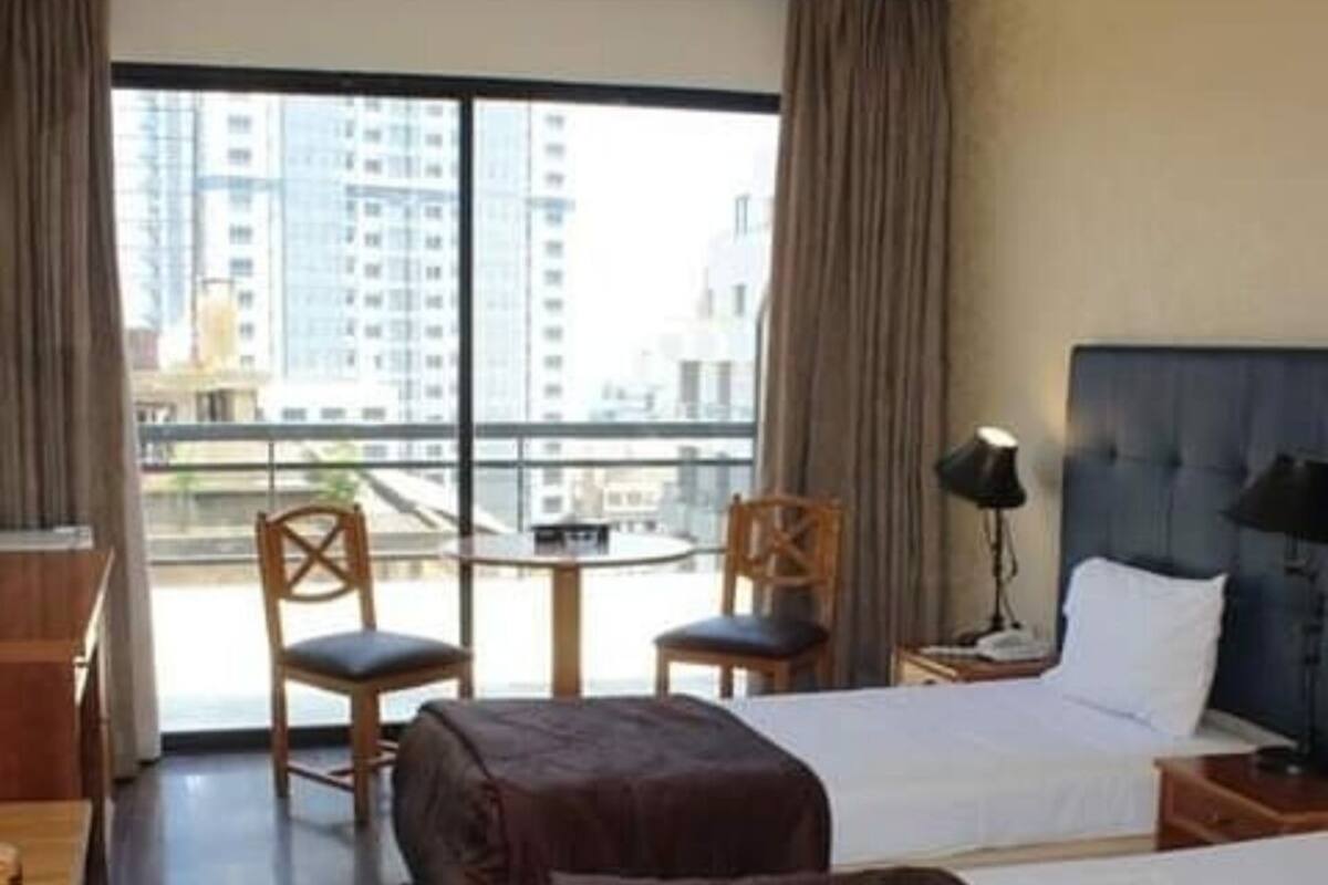 A hotel room with two beds, a desk, a chair, a lamp, and a view of buildings through large windows.