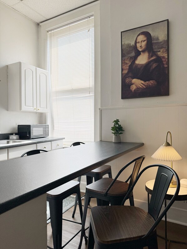 Een moderne keuken met een eethoek, voorzien van een groot raam, witte kastjes, een schilderij van de Mona Lisa en een kleine plant.