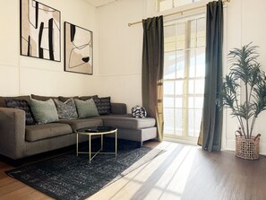 Een moderne woonkamer met een hoekbank, een salontafel en een groot raam met gordijnen.