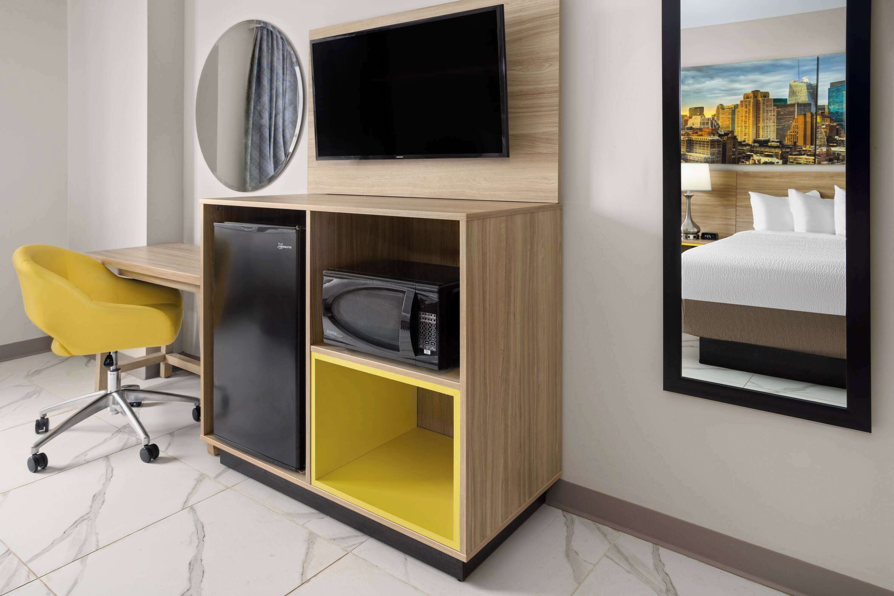 A hotel room with a wooden desk, a flat-screen TV, a microwave, a yellow chair, and a bed with a cityscape view.