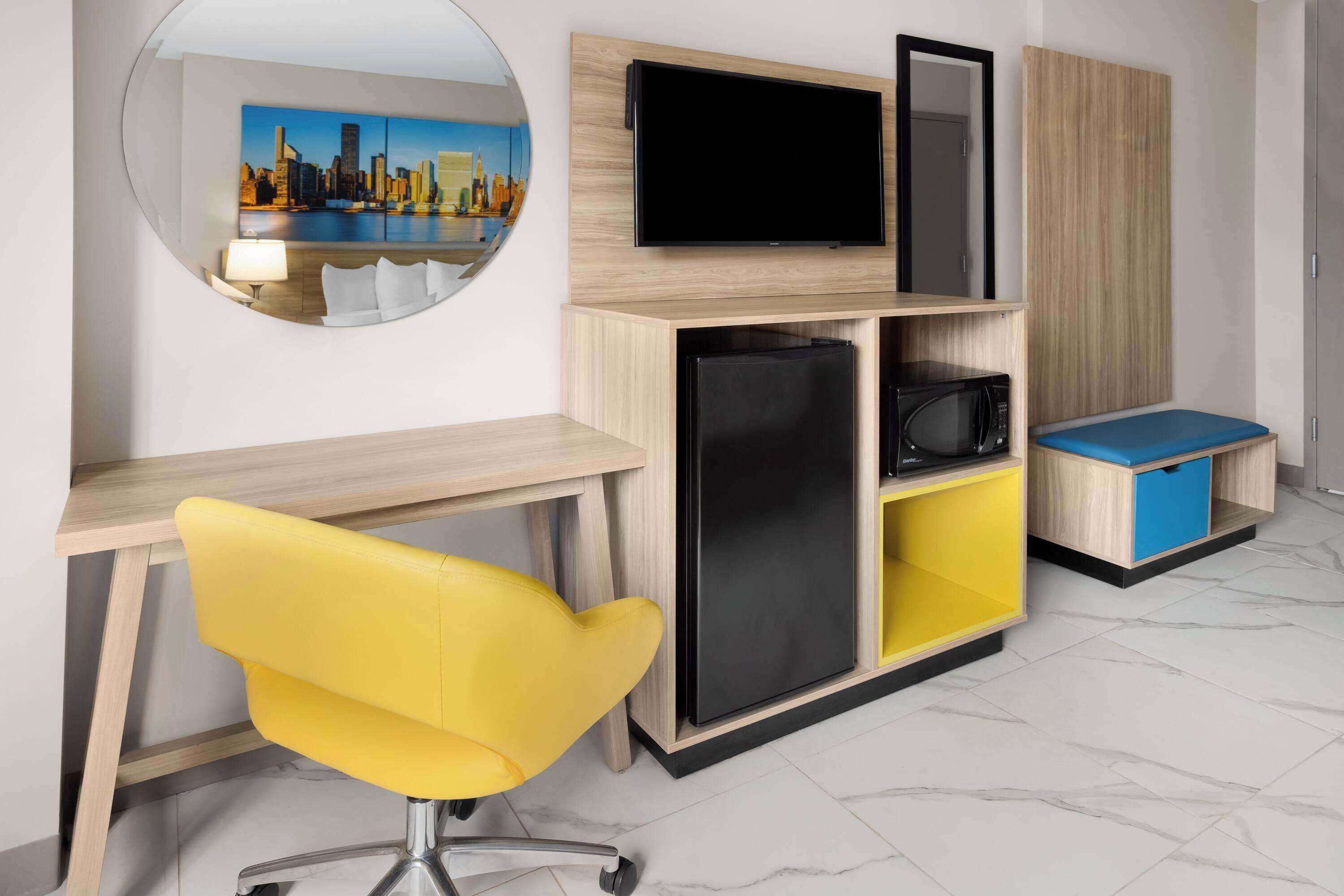 A modern room with a wooden desk, a yellow chair, a flat-screen TV, a microwave, and a refrigerator.