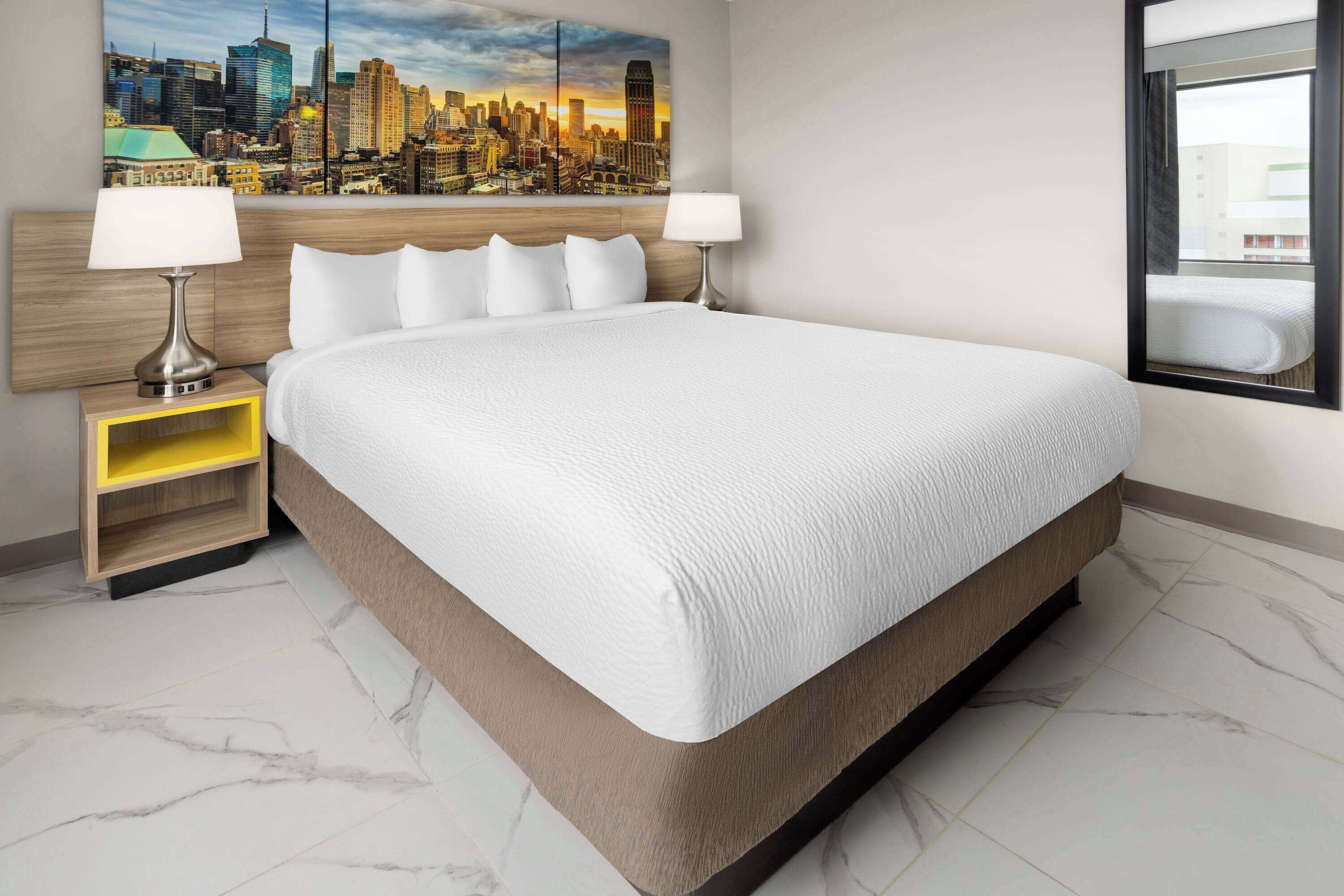 A hotel room with a large bed, two bedside tables with lamps, a cityscape painting on the wall, and a window with a view of buildings.