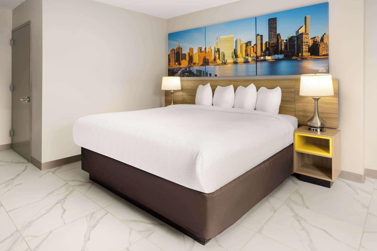 A hotel room with a large bed, two bedside tables with lamps, a cityscape painting on the wall, and a door to the left.