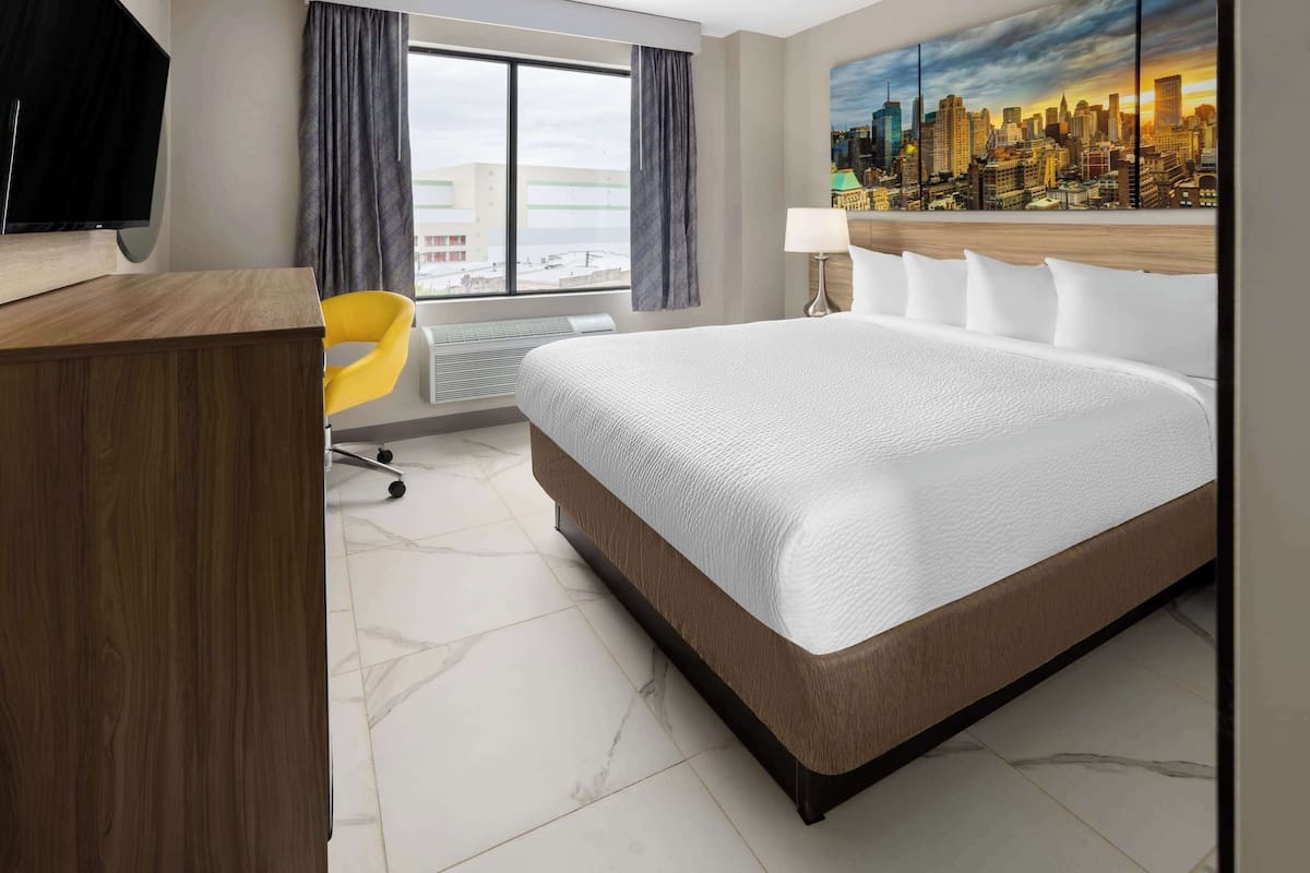 A hotel room with a large bed, a desk, a chair, a TV, a window with curtains, and a cityscape painting on the wall.