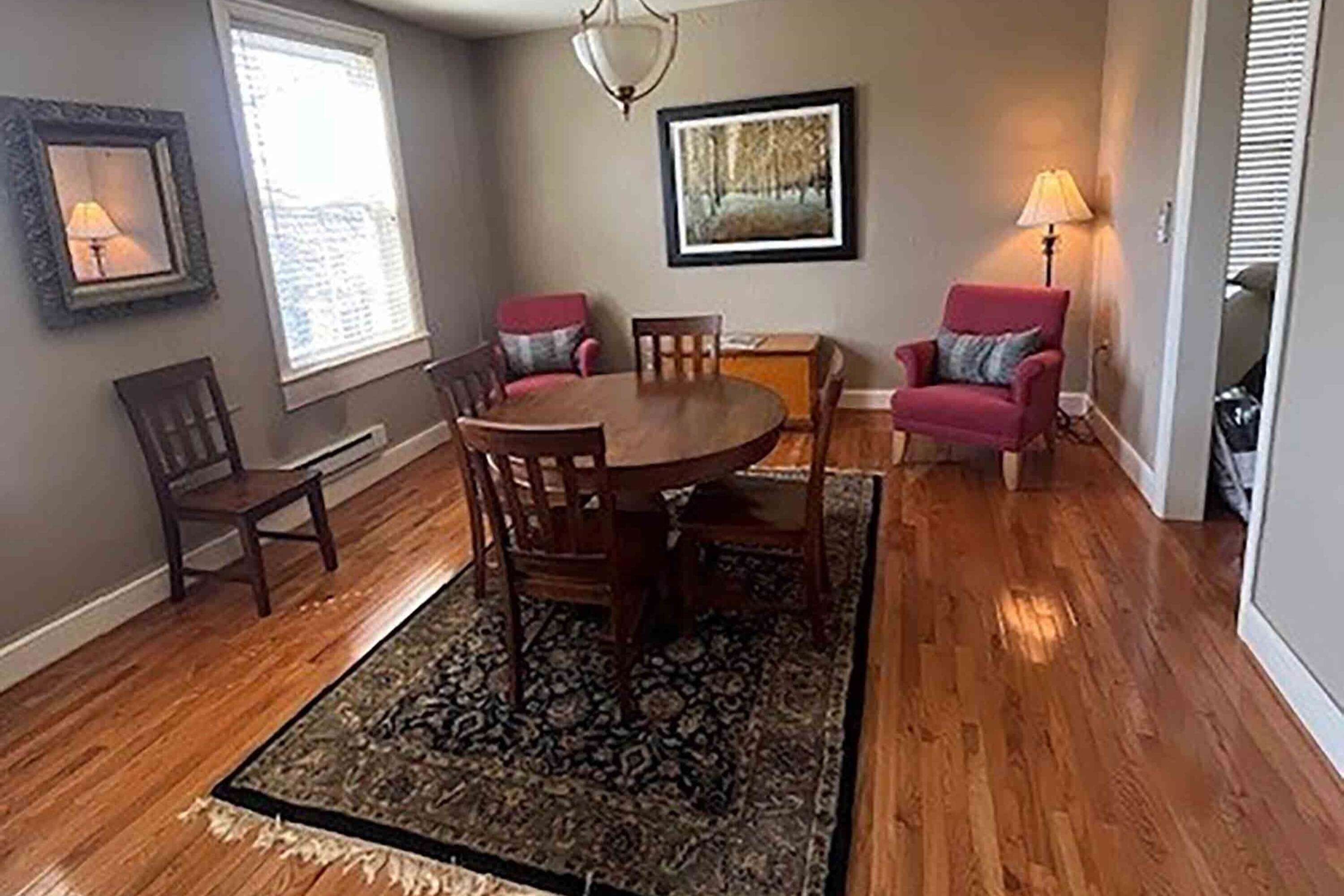 A dining room with a wooden table and chairs, a patterned rug, and two red armchairs.