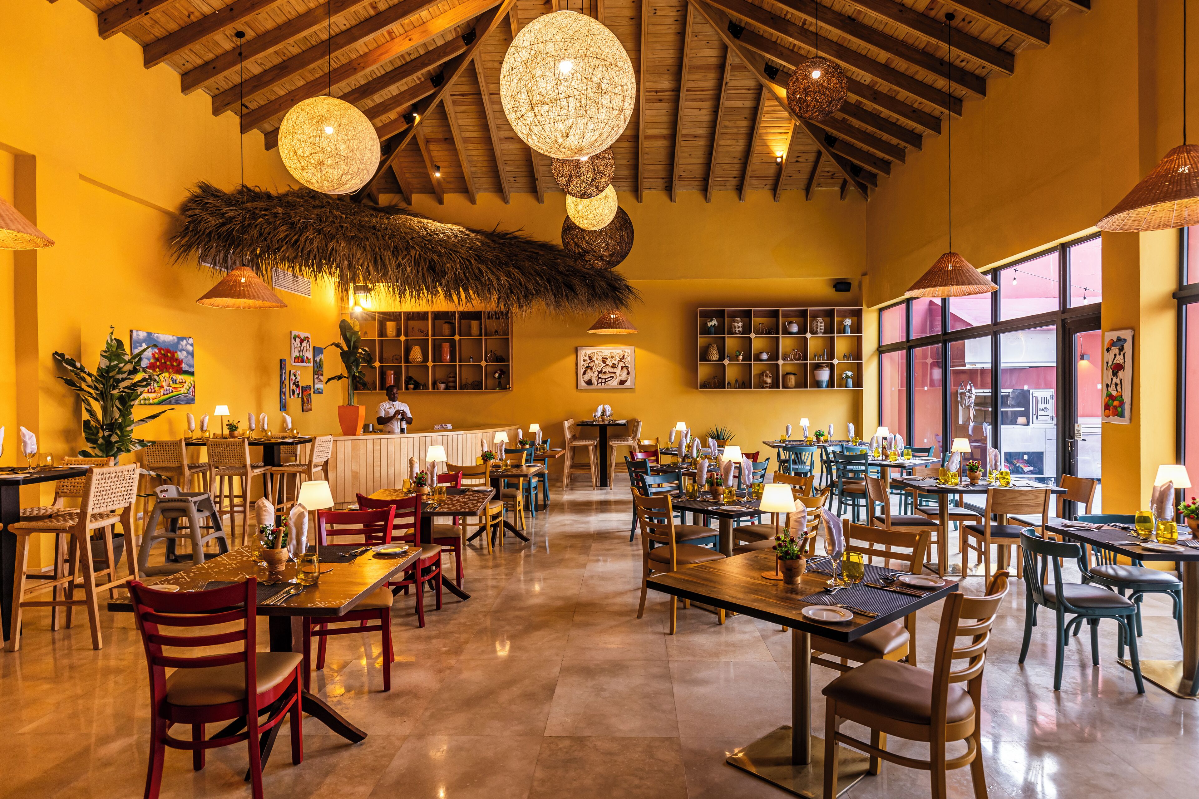 Un restaurant spacieux avec de hauts plafonds, des poutres en bois et des luminaires sphériques suspendus. Les tables sont accompagnées de chaises, et les murs sont ornés d’œuvres d’art encadrées et d’étagères où sont exposés des objets décoratifs.