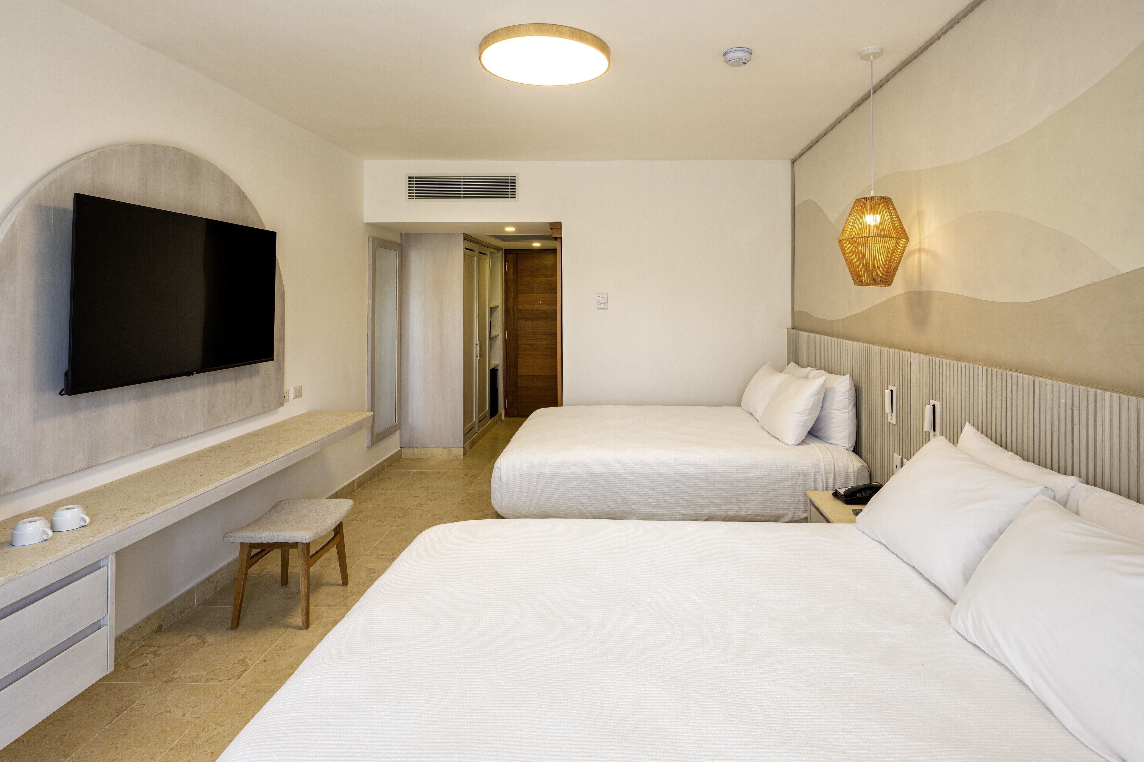 A hotel room with two beds, a TV mounted on the wall, a bench, and a bedside table with a lamp.