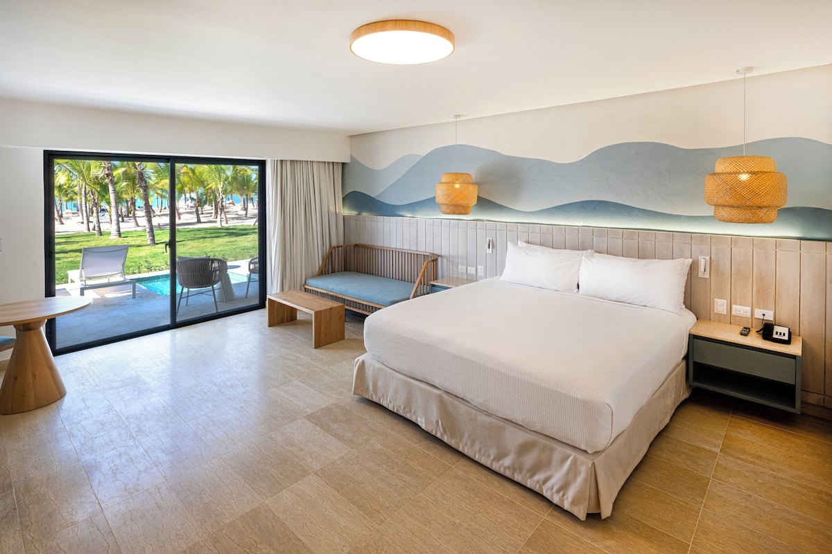Une chambre d’hôtel moderne dotée d’un grand lit, d’un coin salon, d’une table à manger et offrant une vue sur la plage et les palmiers, visible à travers la porte vitrée.