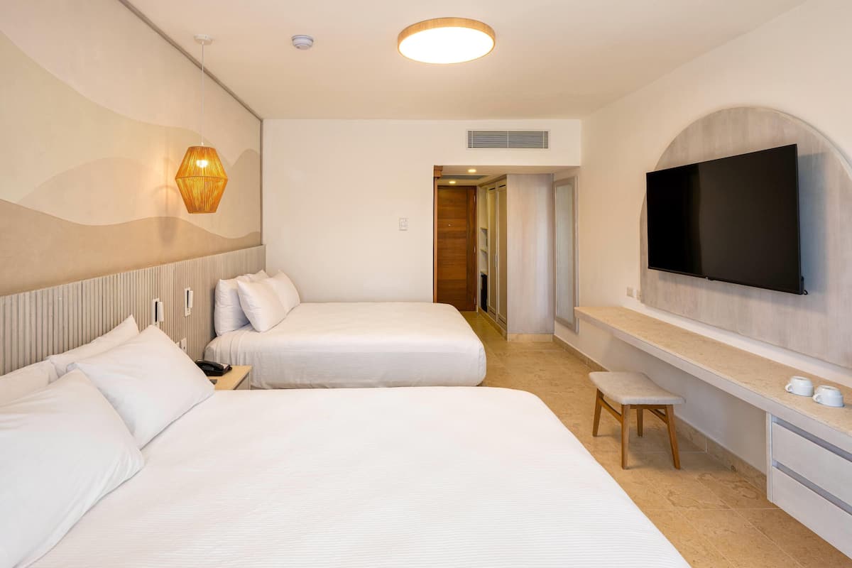 Une chambre d’hôtel avec deux lits, un téléviseur fixé au mur, une table de chevet avec une lampe et un petit tabouret.