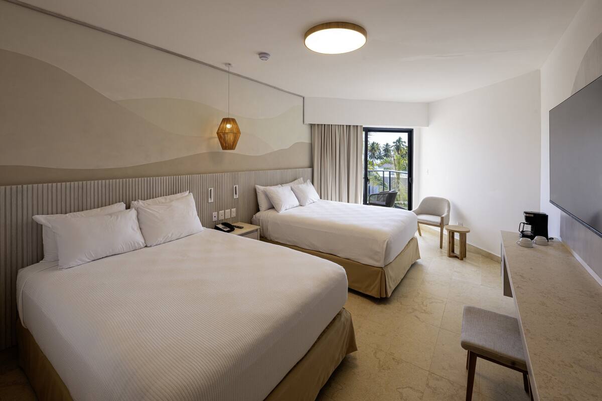 Une chambre d’hôtel avec deux lits, un bureau, une chaise, une télévision et un balcon offrant une vue sur les palmiers.