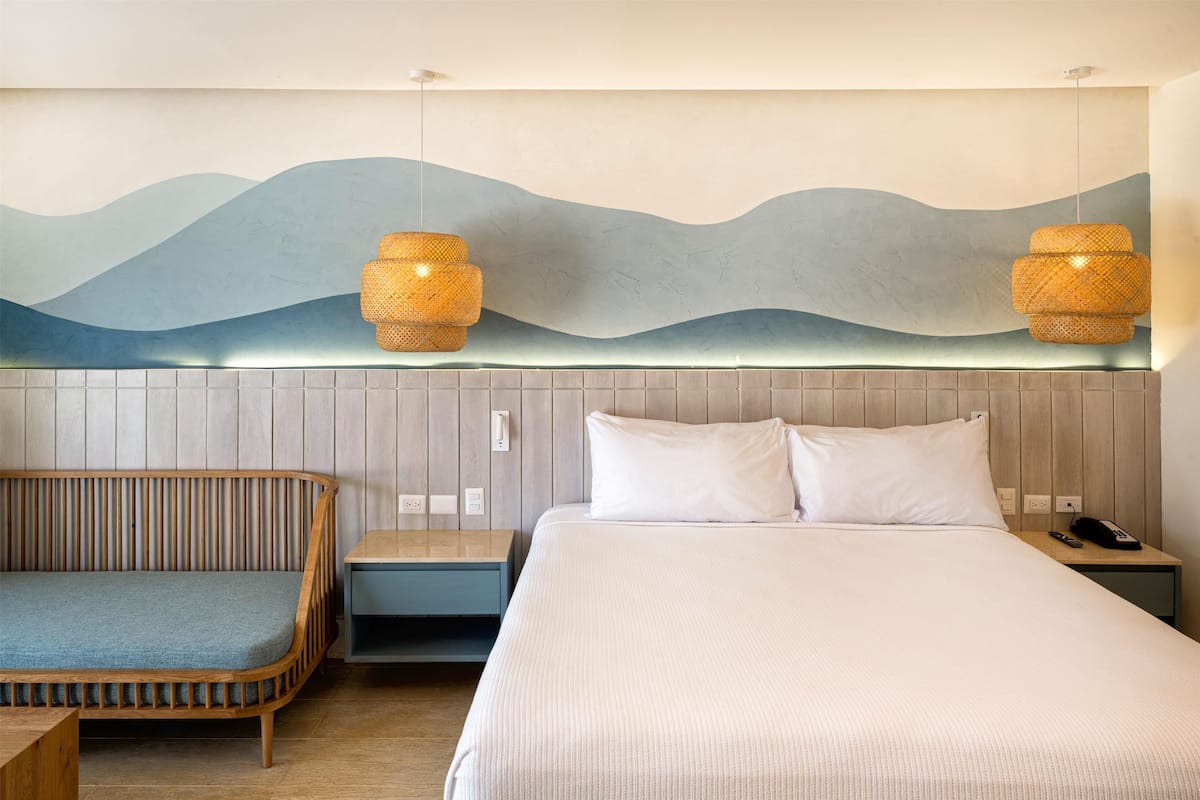 Une chambre d’hôtel comprenant un lit, une banquette, une œuvre d’art accrochée au mur et deux lampes suspendues.