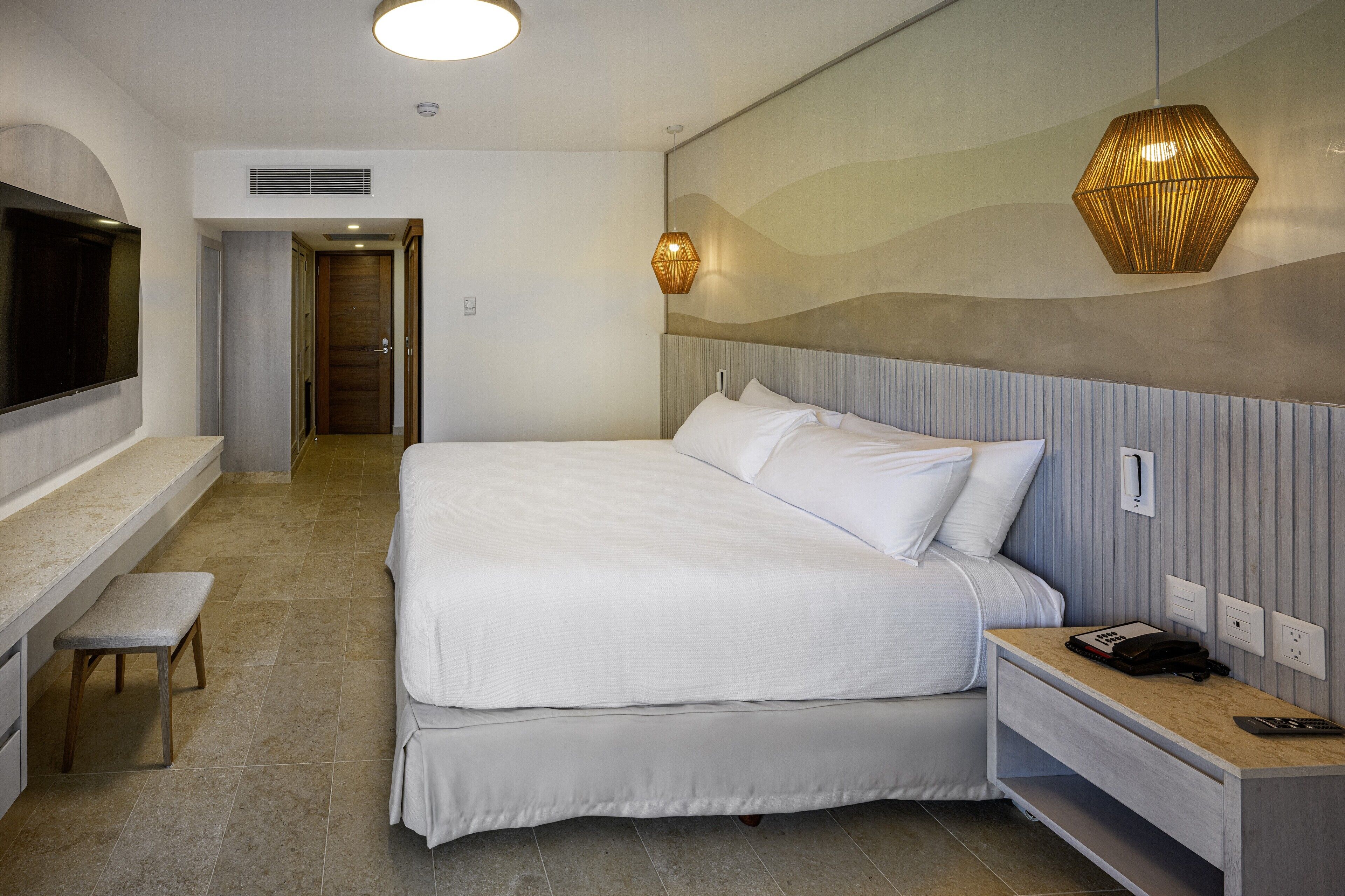A modern hotel room with a large bed, a bedside table with a phone, a bench, and a TV mounted on the wall.