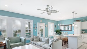 A modern kitchen with white cabinetry, a dining area with a white table and chairs, and a living room with a light blue sofa and a ceiling fan.