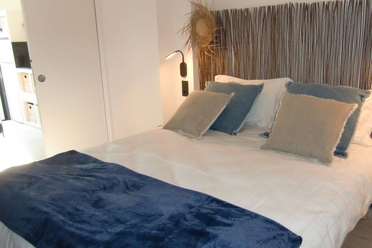 Et soveværelse med en seng, puder, et natbord, en lampe og en væg med lodrette linjer.