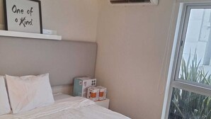 A bedroom with a bed, a bedside table with boxes, a wall-mounted air conditioner, a window with a view, and a sign on the wall. - Gorgeous 2BDRM with A/C - Miraflores (Miraflores)