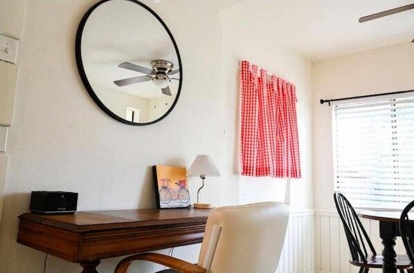 Una habitación con un escritorio de madera, una silla, un ventilador de techo, una ventana con persianas y una cortina a cuadros rojos y blancos.