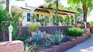 A yellow building with a covered porch, surrounded by a garden with various plants and a brick wall. - Farmhouse Escape w/Mountain Views (Tolleson)