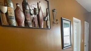 A hallway with a long wooden bench and stools, a wall-mounted sculpture of various bottles, and a framed picture.