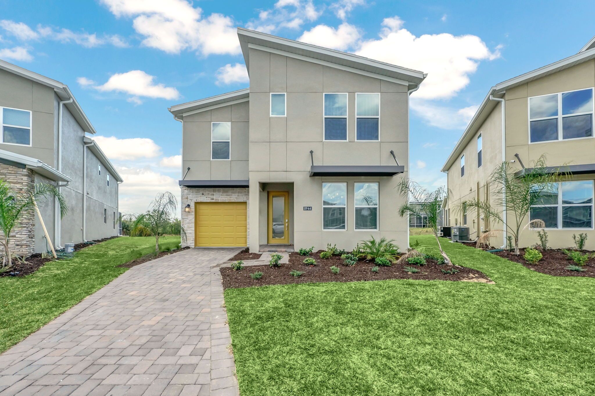 A modern two-story residential building with a yellow garage door, a brick pathway leading to the entrance, and well-maintained landscaping.