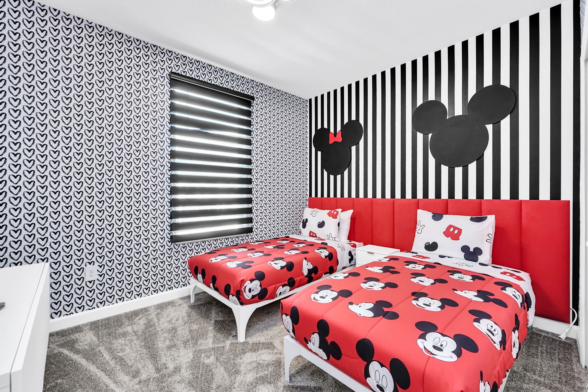 A room with two beds, each with red bedding featuring Mickey Mouse prints, a red upholstered headboard, and a wall with Mickey Mouse silhouettes.
