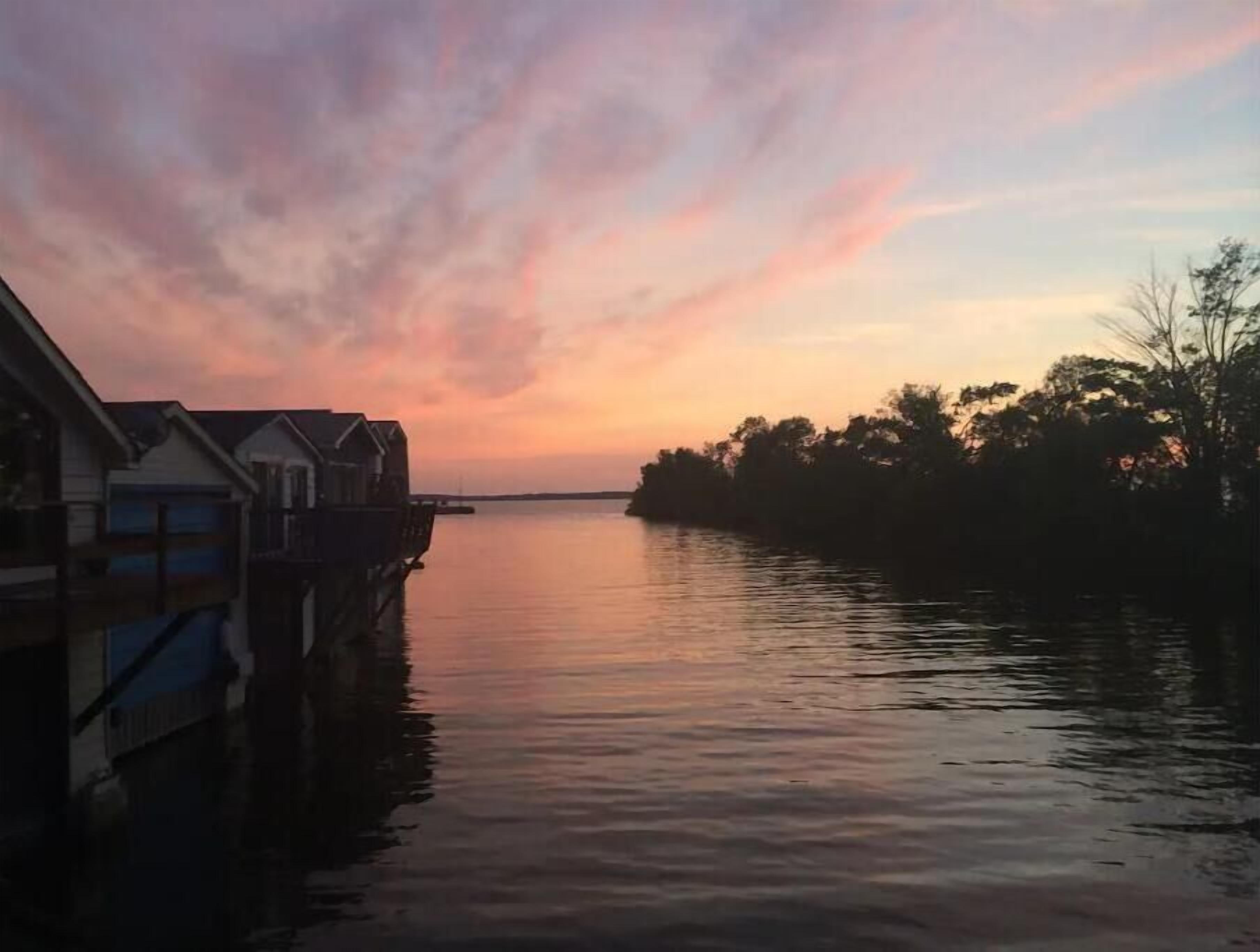 Une propriété située au bord de l’eau, dotée d’un quai s’étendant dans une eau calme, entourée d’arbres et offrant une vue sur un ciel au coucher du soleil.