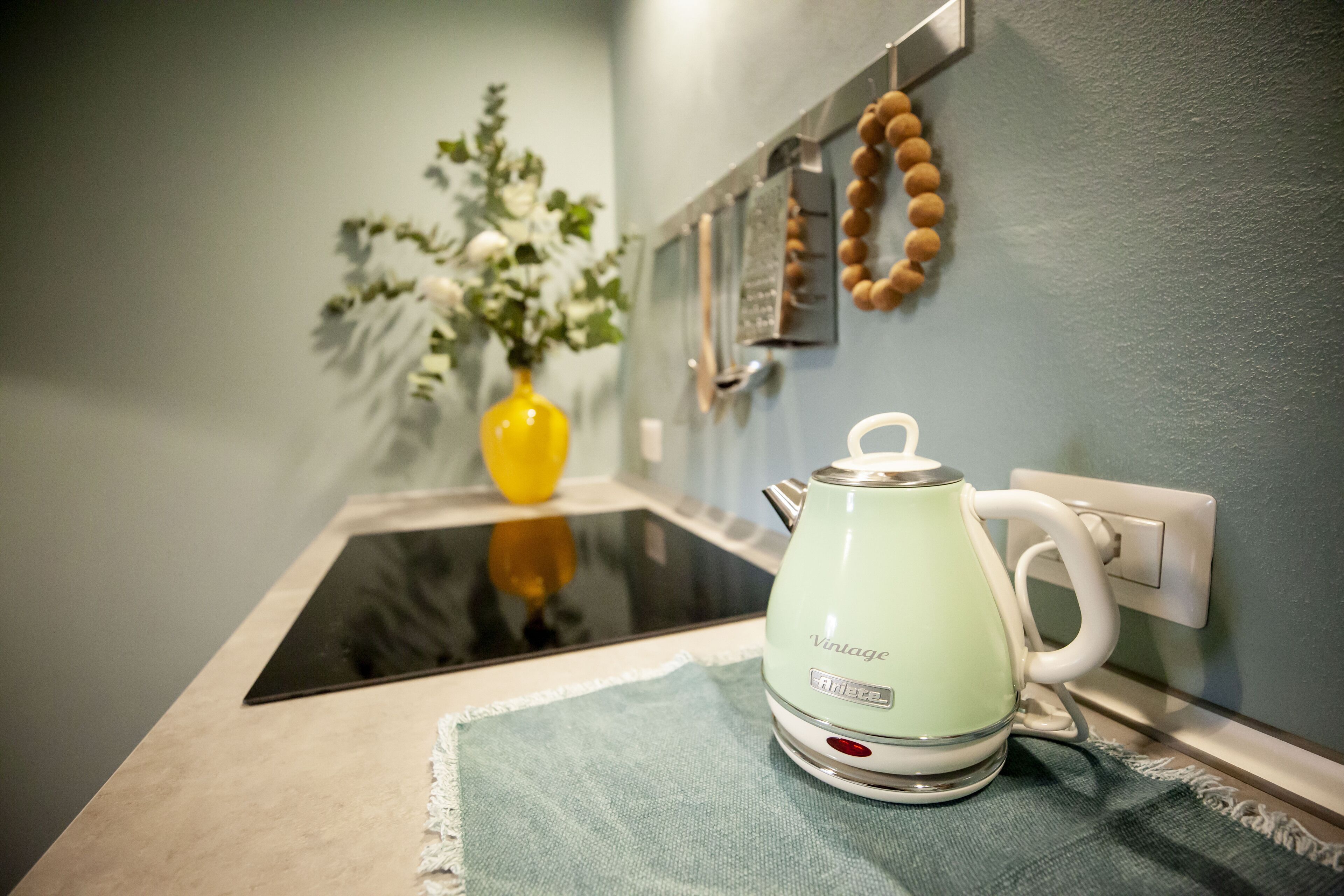 Een keukentafel met een groene waterkoker, een gele vaas met witte bloemen en een aan de muur bevestigde thermometer.